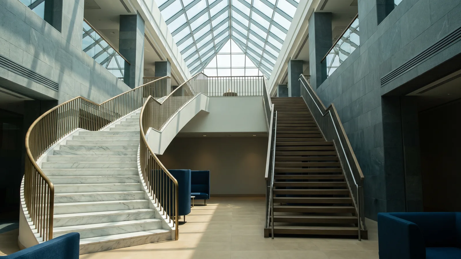 Two staircases in a bright office atrium leading to the same upper floor: a wide sweeping marble staircase and a plain straight wooden one
