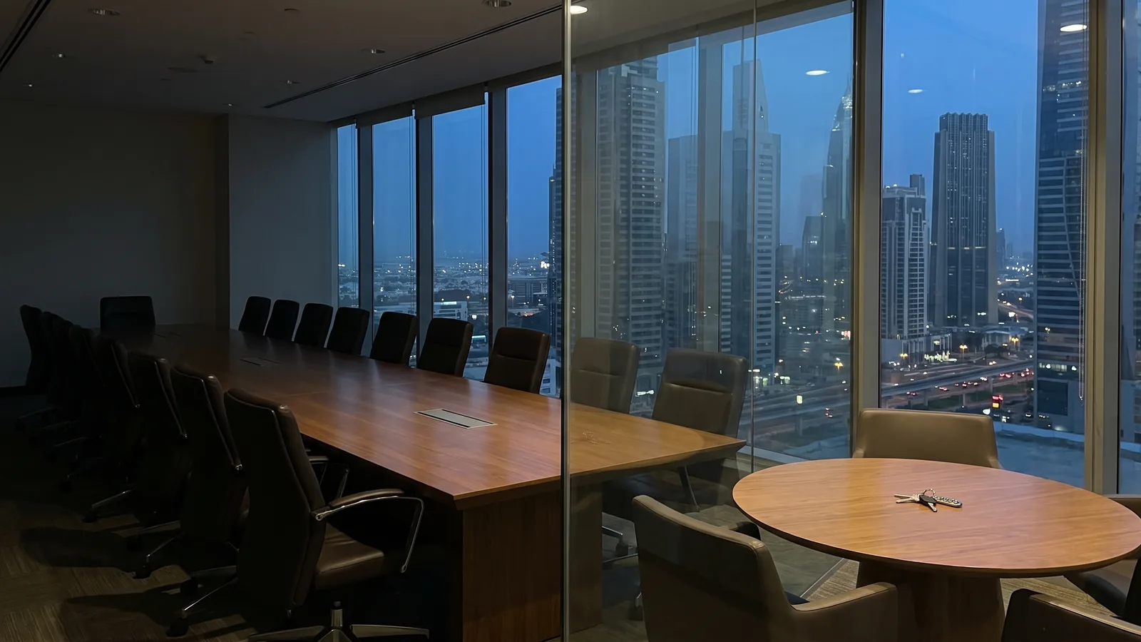 A long empty boardroom table beside a small round two-seat table with a set of keys on it, in a Dubai office tower at dusk