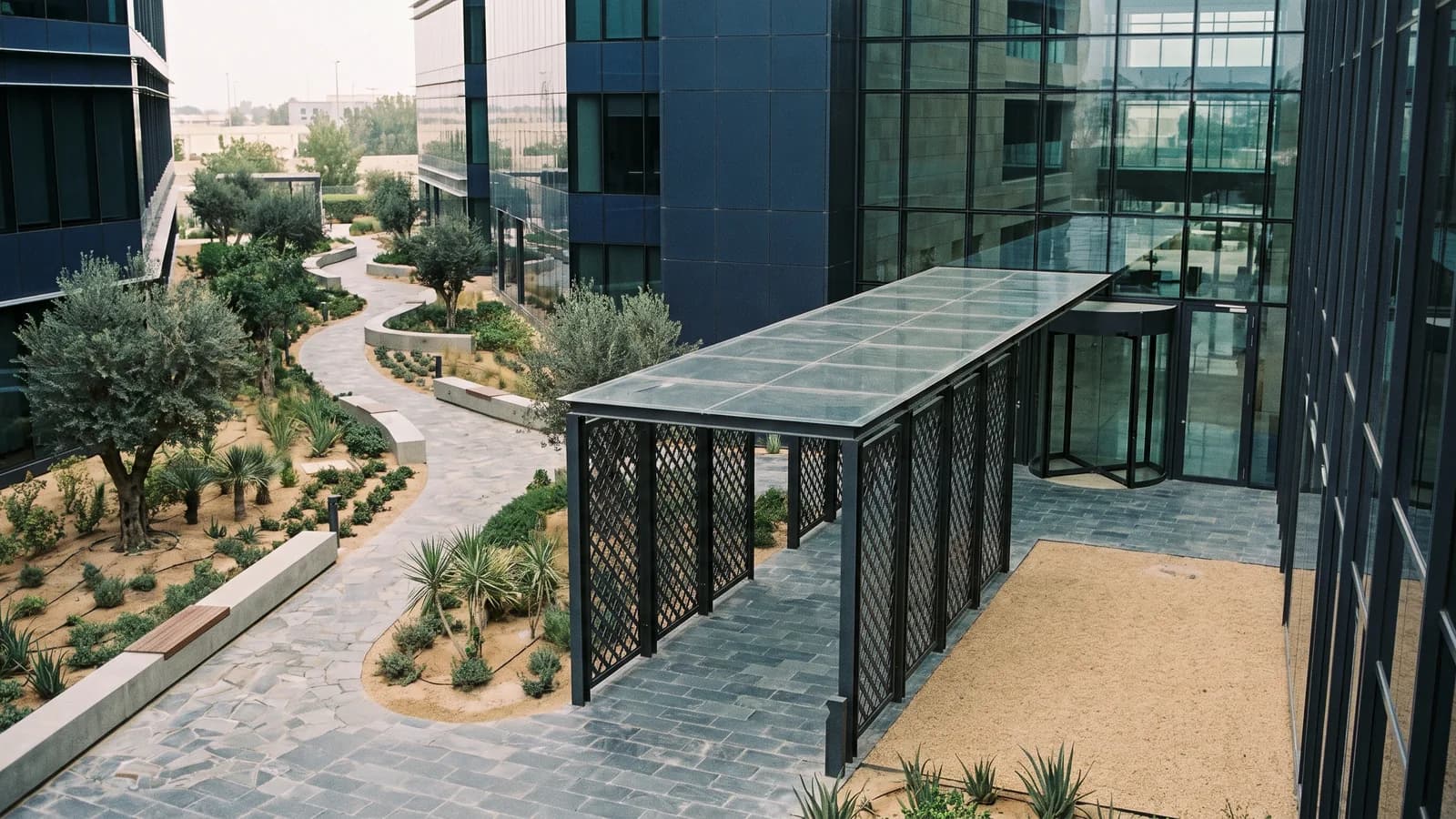 A courtyard between glass office buildings where a winding garden path and a straight covered walkway both lead to the same entrance