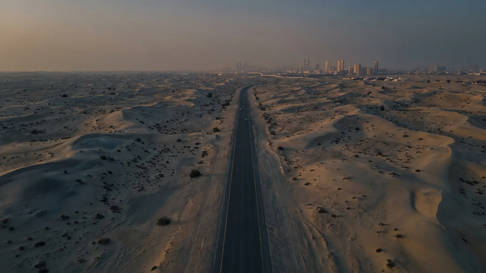 An aerial view at golden hour of the desert highway between Dubai and Abu Dhabi, dunes on either side and a city skyline on the horizon