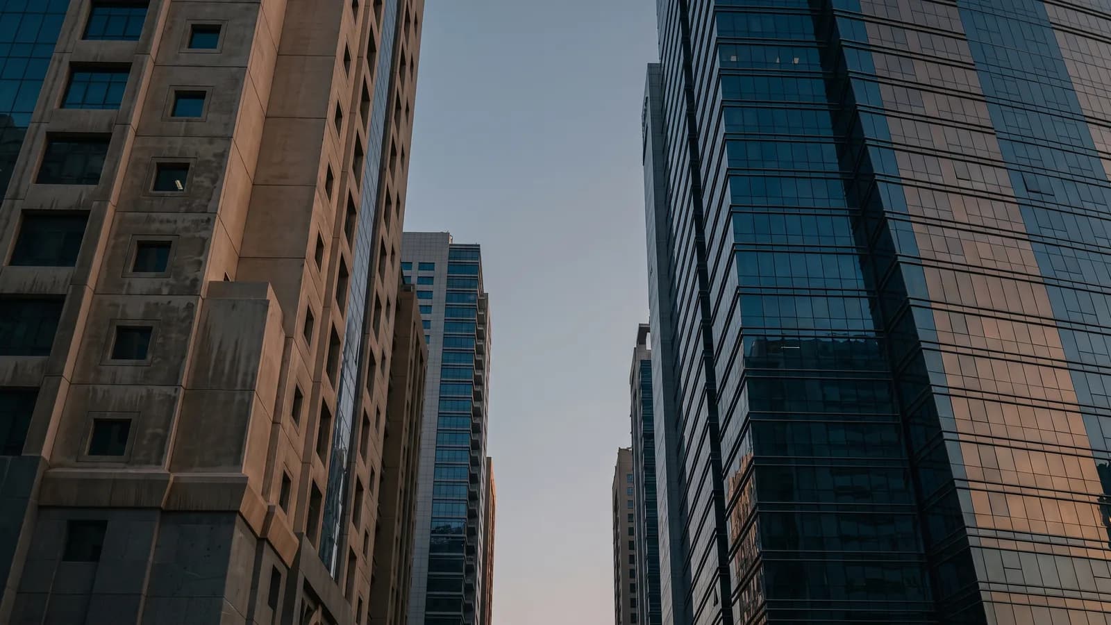 An older stone-clad office tower and a newer glass tower standing side by side in Dubai, seen from street level in early evening light