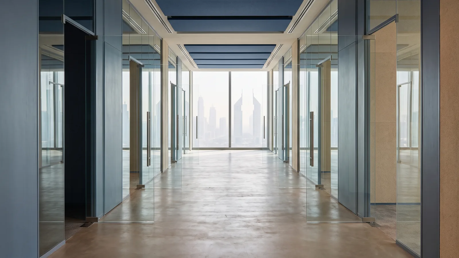 A long bright corridor in a modern Dubai office lined with open glass doors leading toward a window full of daylight