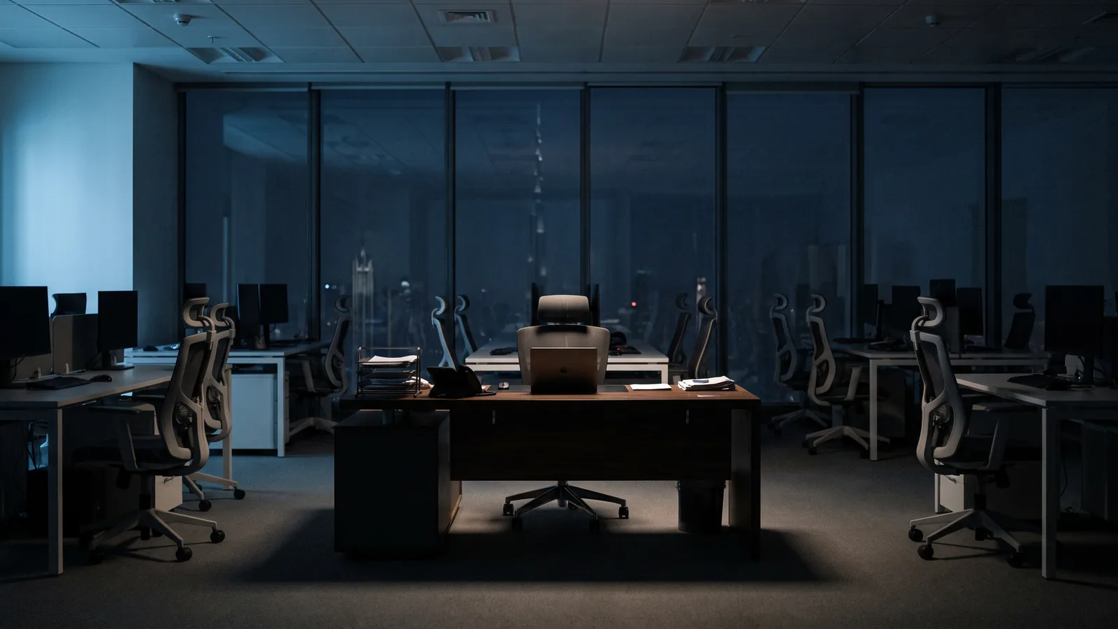 A dark modern office at night where a single desk is lit and the rest sit in shadow, with the Dubai skyline through the glass wall